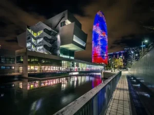 Paisaje urbano nocturno de Barcelona con vista de la torre Agbar y un edificio iluminado con un jardín con agua delante